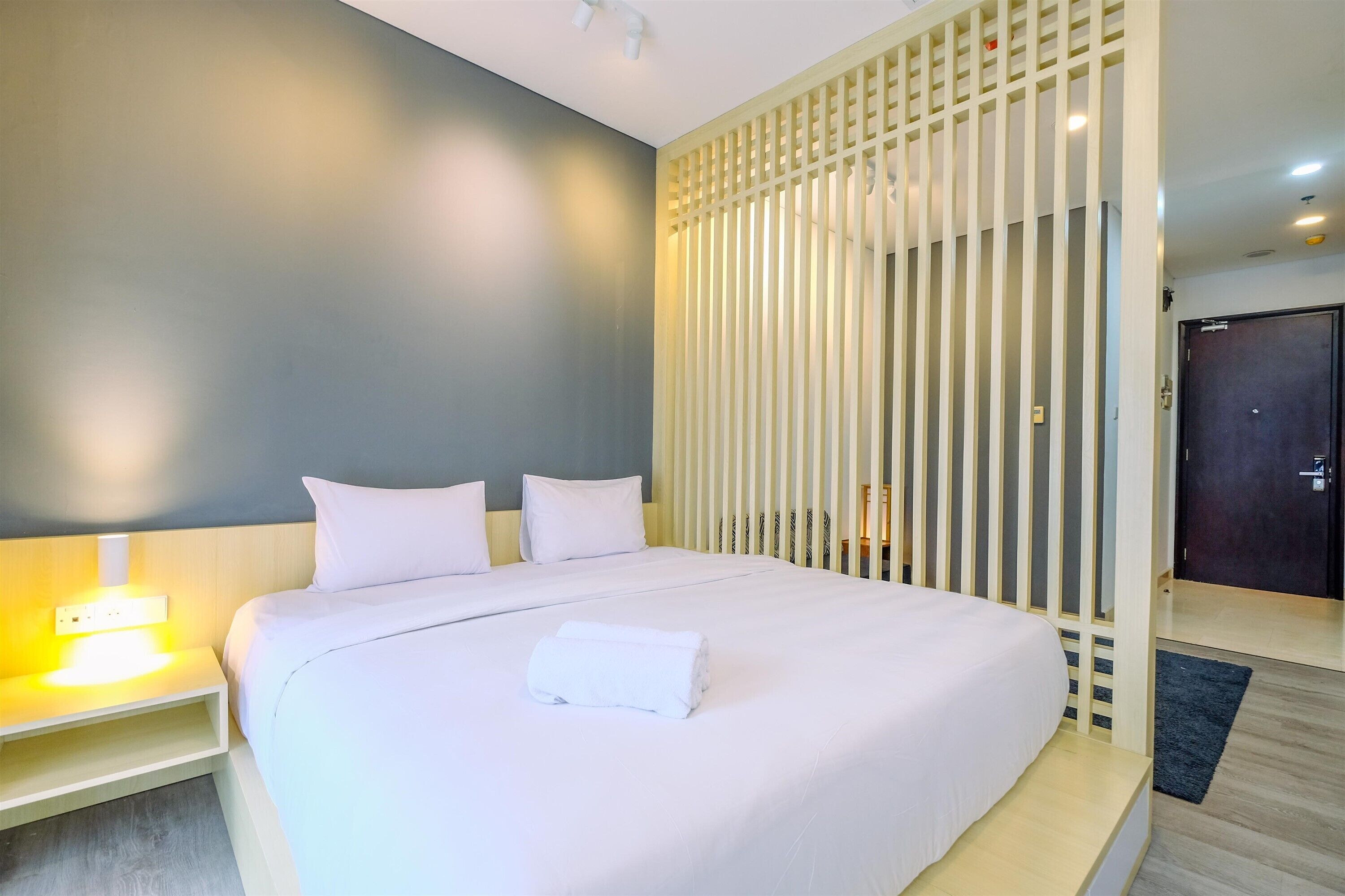 Un lit bien fait, avec du linge de lit blanc et une serviette enroulée, une table de chevet avec une lampe et un plancher en bois.