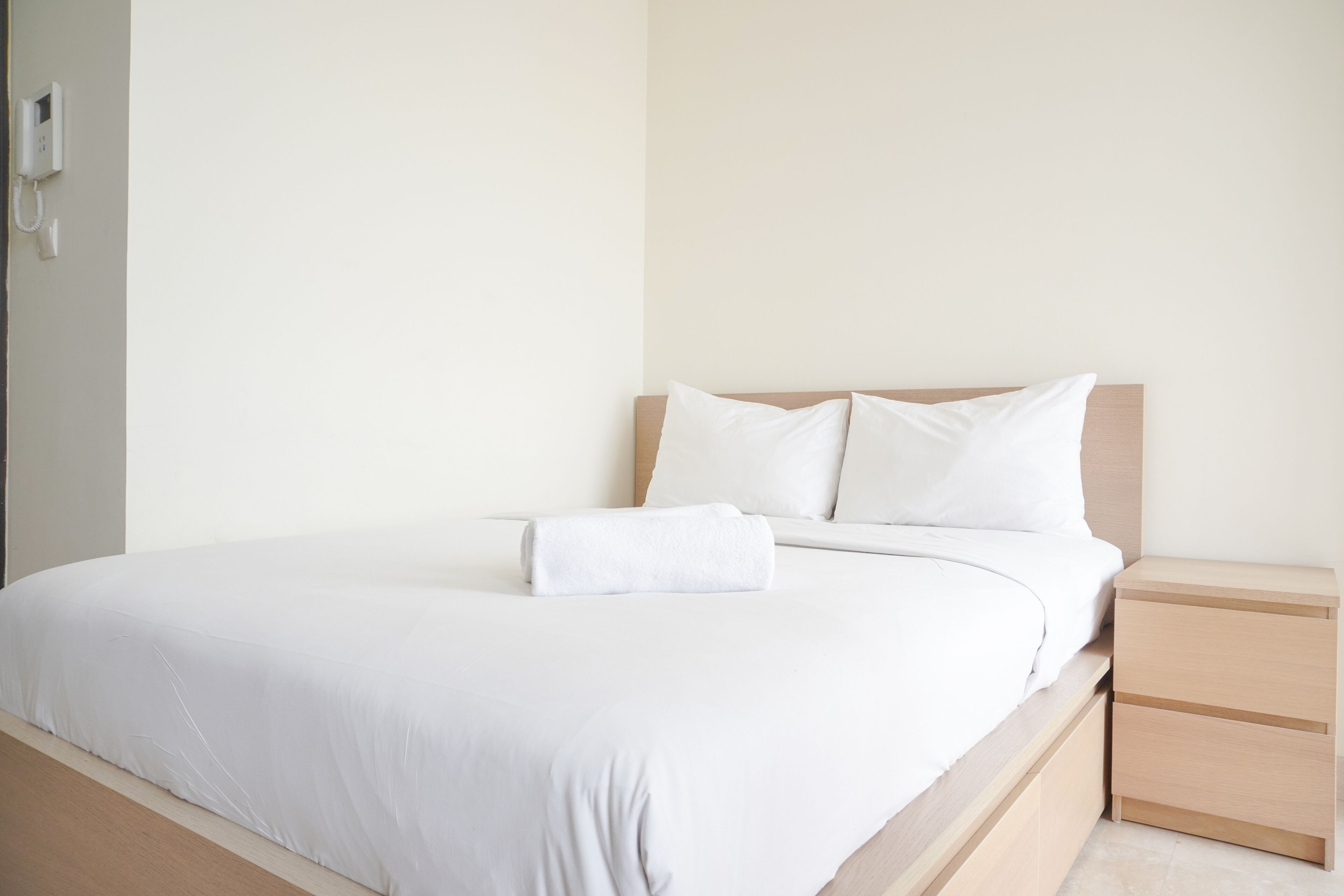 A neatly made bed with white linens and pillows, a wooden headboard, and a small bedside table with drawers.