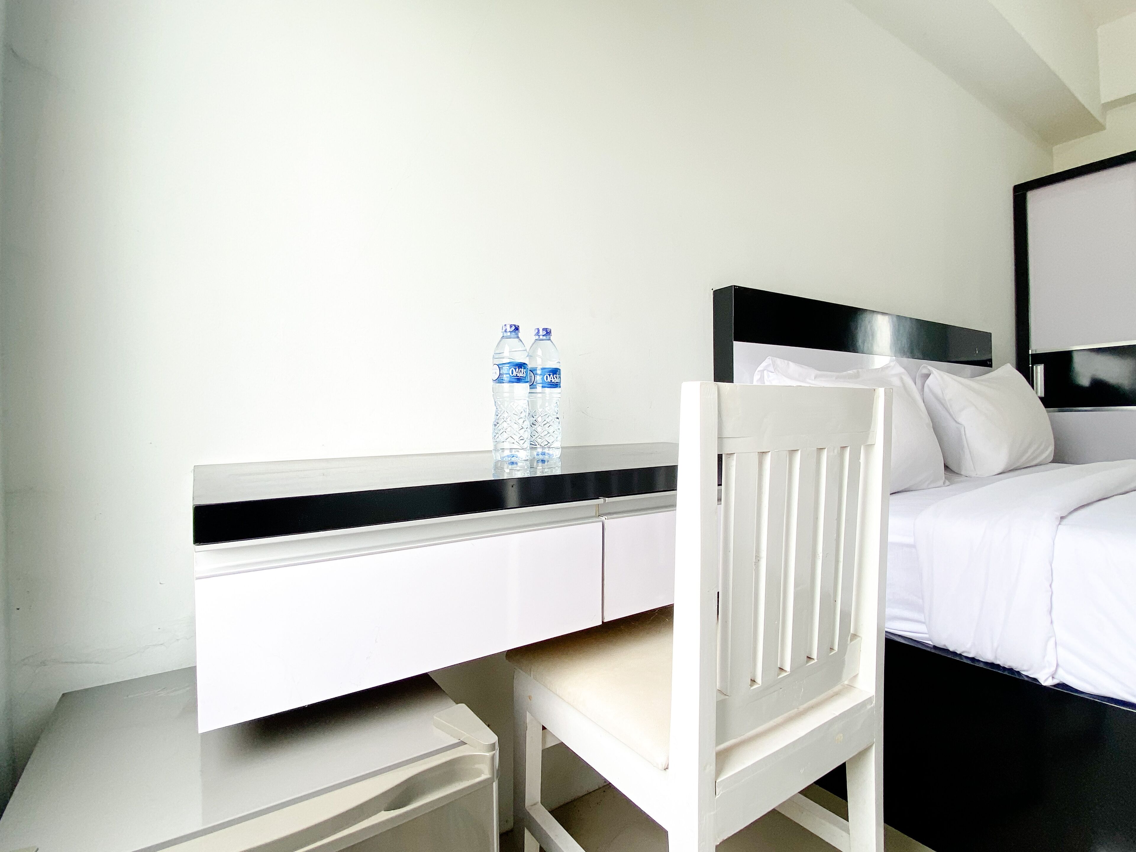 Habitación de hotel con un escritorio blanco, una silla, una cama con ropa blanca y un cabecero negro. Hay dos botellas de agua sobre el escritorio.