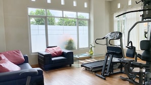 A home gym with a treadmill, weight bench, and exercise equipment. There is a large window with a view of trees and a white fence. A chair with a pink cushion is also visible. - Simple And Homey 2Br At Dian Regency Apartment (Surabaya)