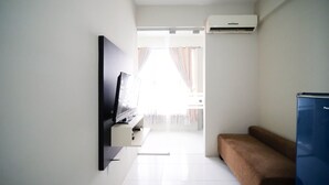 A hotel room with a flat-screen TV mounted on the wall, a blue refrigerator, and a brown sofa. - Simple And Homey 2Br At Dian Regency Apartment (Surabaya)