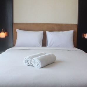 A neatly made bed with white linens and two rolled towels, a wooden headboard, and two wall-mounted lamps. - Warm 1Br Apartment (No Kitchen) At Marbella Suites Dago Pakar Bandung (Bandung)
