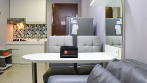 A compact kitchen with white cabinets, a tiled backsplash, and a refrigerator with papers on it. There's a table with a bag labeled 'Travelio' and a couch in the foreground. - Minimalist And Warm 1Br Signature Park Grande Apartment (Jakarta)