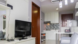 A modern living room with a flat-screen TV, a white sofa, and a kitchen area with white cabinets and a tiled backsplash. - Minimalist And Warm 1Br Signature Park Grande Apartment (Jakarta)