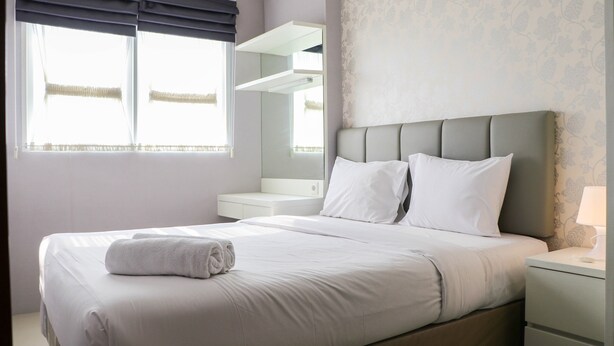 A neatly made bed with white linens and a folded towel, a bedside table with a lamp, and a large mirror above the bed. - Minimalist And Warm 1Br Signature Park Grande Apartment (Jakarta)
