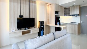 A modern living room with a flat-screen TV, a sofa, and a kitchen area with a microwave and refrigerator. - Spacious And Homey 1Br Vasanta Innopark Apartment (Cikarang)