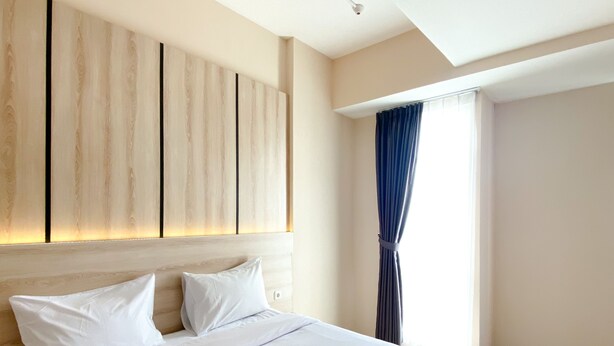 A hotel room with a large bed, a headboard with vertical light panels, a window with curtains, and a view of the outside. - Spacious And Homey 1Br Vasanta Innopark Apartment (Cikarang)