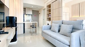 A modern living room with a grey sofa, a flat-screen TV, and a kitchen area with a washing machine. - Spacious And Homey 1Br Vasanta Innopark Apartment (Cikarang)