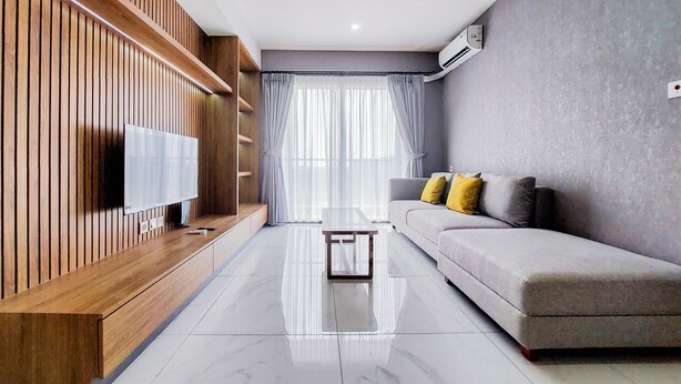 A modern living room with a wooden accent wall, a grey sofa, a glass coffee table, and a flat-screen TV. - Comfort And Spacious 3Br At Sky House Bsd Apartment (Pagedangan)