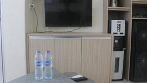 Ein Zimmer mit flachbildfernseher an der Wand, einer Kaffeemaschine und einem Kühlschrank. Auf einem kleinen Tisch stehen zwei Wasserflaschen.