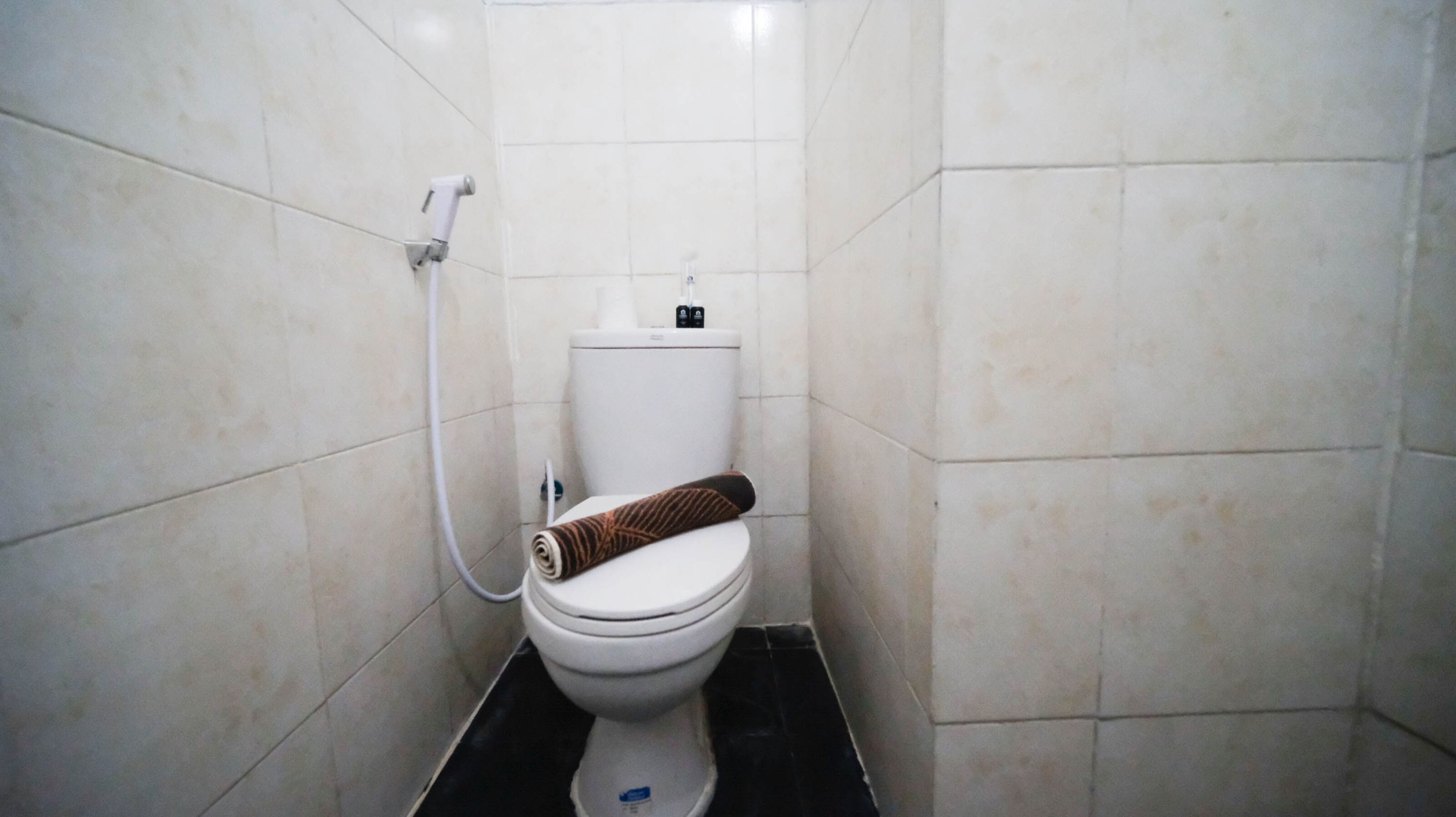 A small, tiled bathroom with a toilet, a white toilet paper holder, and a visible pipe.