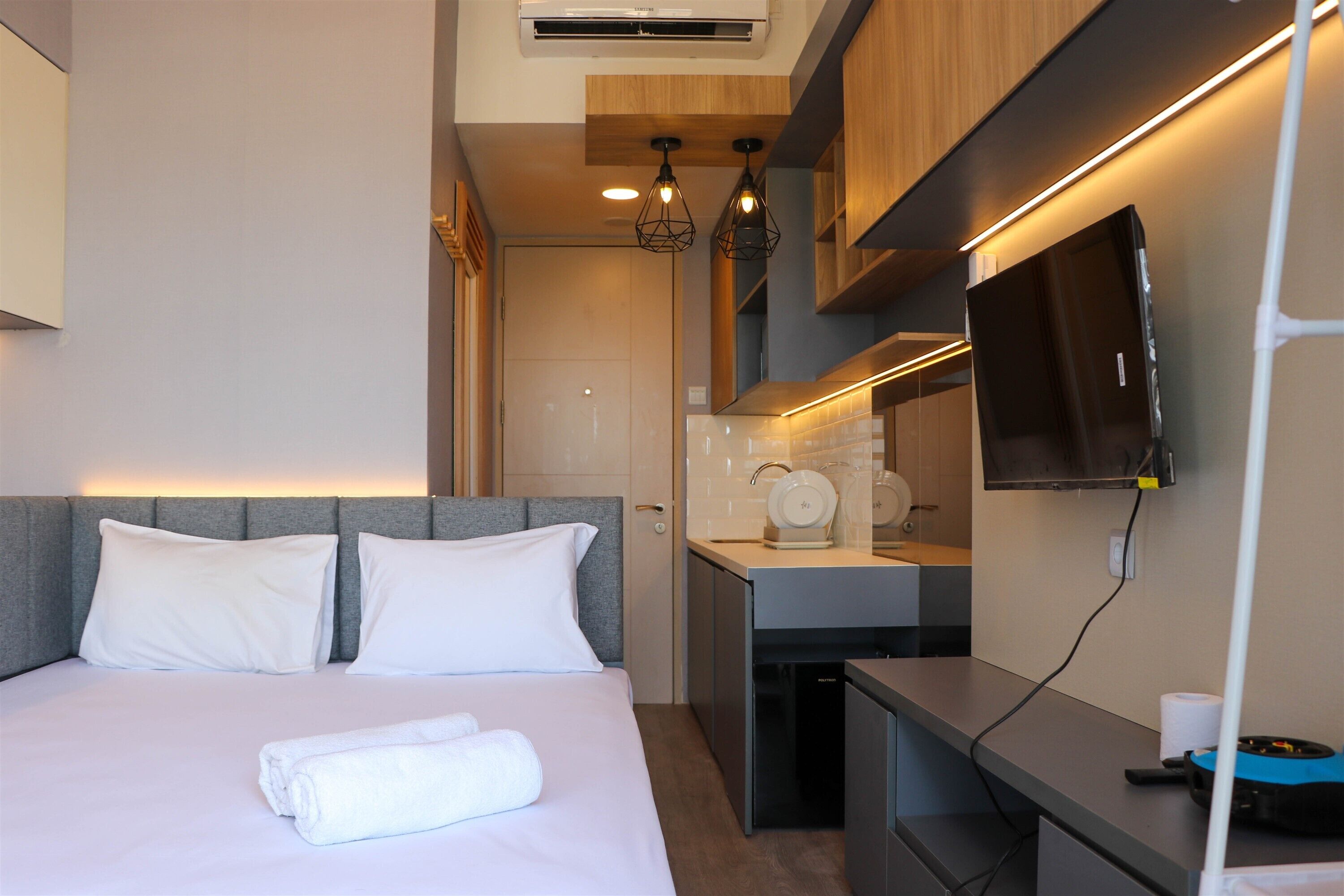 A modern hotel room with a bed, a small kitchenette, a flat-screen TV, and a ceiling-mounted air conditioner.