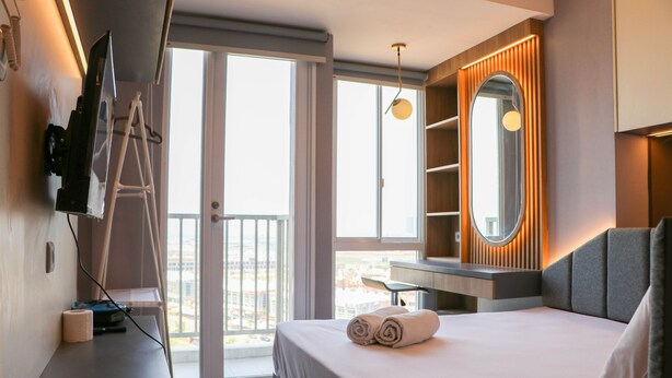 Quarto de hotel moderno com uma cama grande, uma TV fixada na parede, uma mesa com cadeira e uma varanda com vista para a cidade.