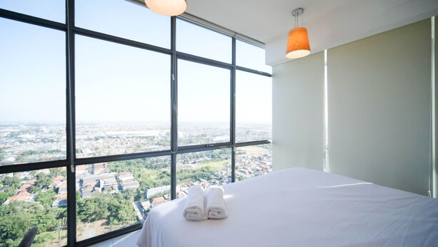 A hotel room with a large window offering a city view, a bed with white linens, and a hanging orange pendant light.