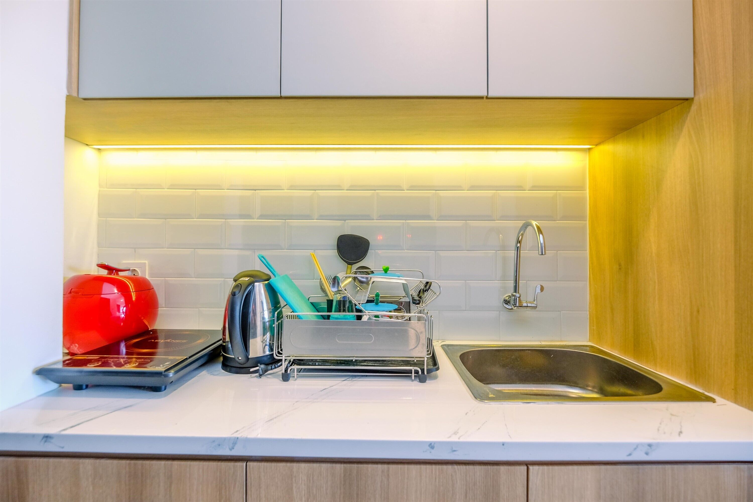 A kitchen with a stainless steel sink, a red kettle, and a dish rack with utensils.