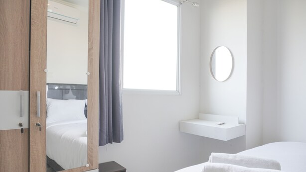 Una habitación de hotel con un armario de madera, una mesita de noche, una cama blanca con toallas blancas, una ventana con cortinas y un lavamanos pequeño y blanco.