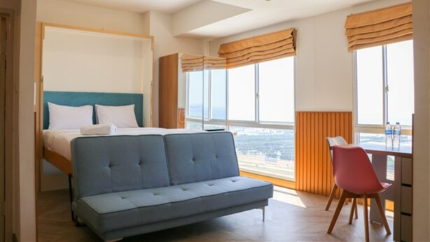 A modern hotel room with a bed, a sofa, a dining table, and large windows offering a view. - Spacious And Cozy Studio Tokyo Riverside Pik 2 Apartment (Jakarta)