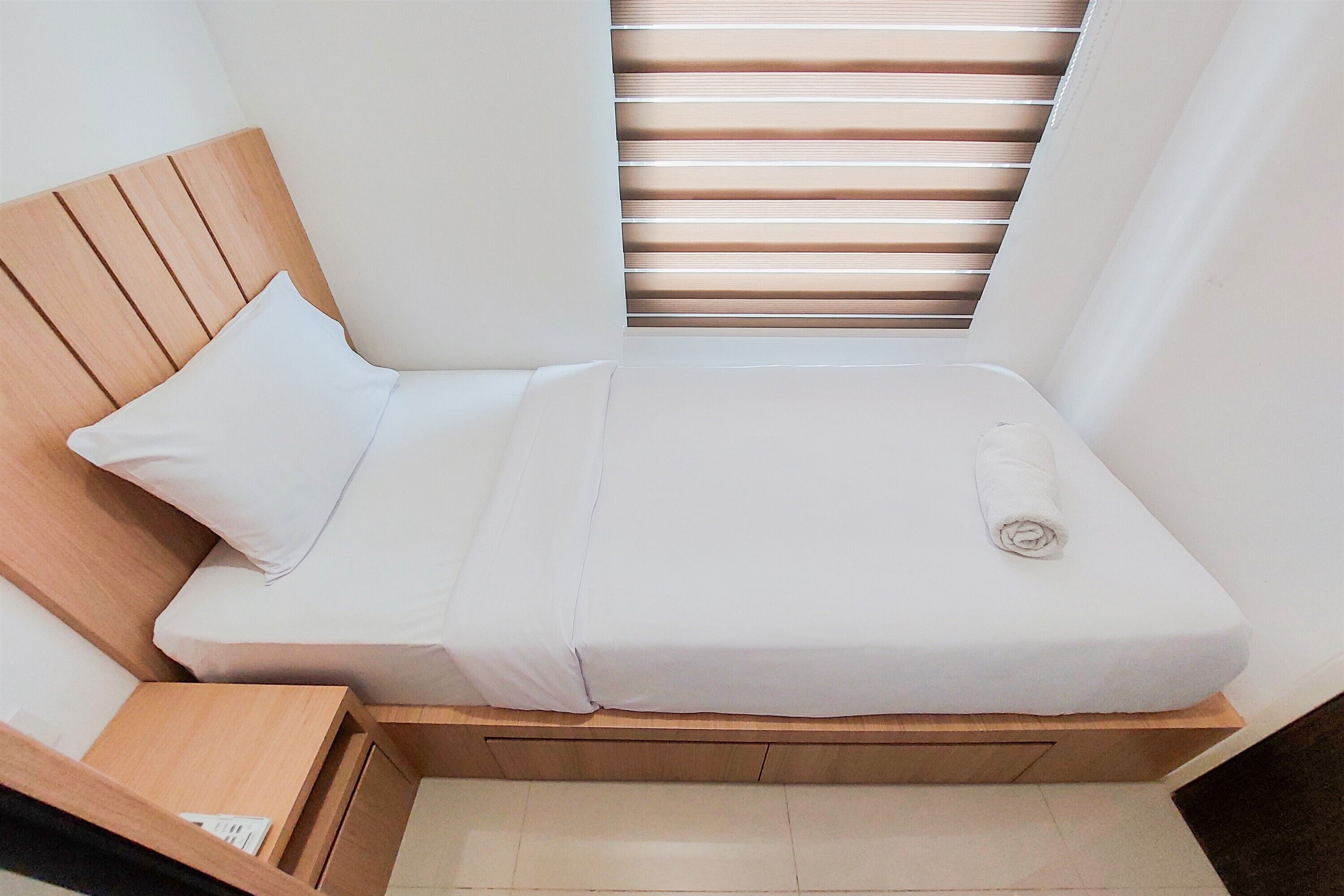A small, neatly made bed with white bedding and a wooden headboard, positioned under a window with horizontal blinds.