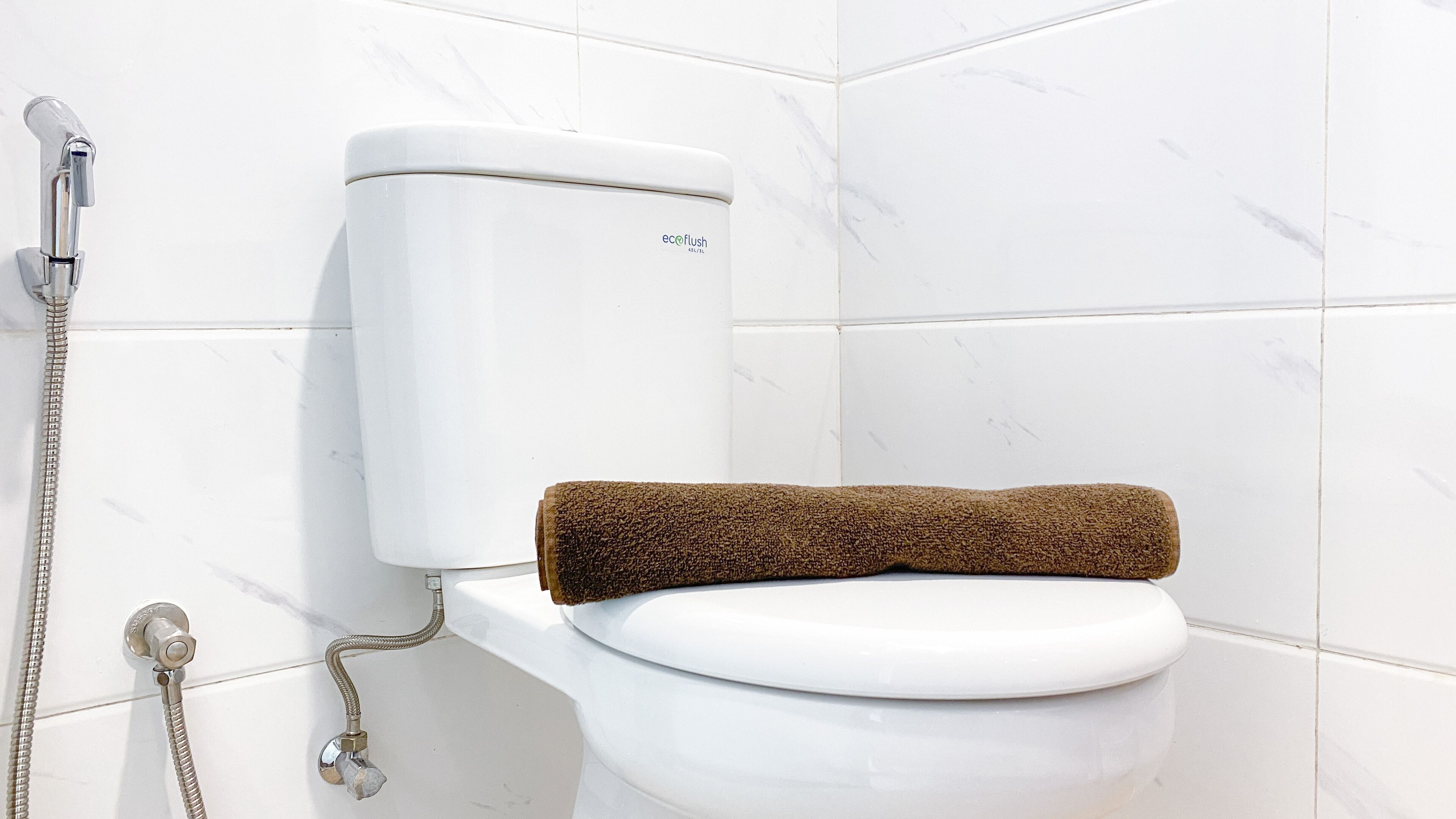 A white toilet with a brown towel on the seat, a silver shower hose, and a white toilet tank.