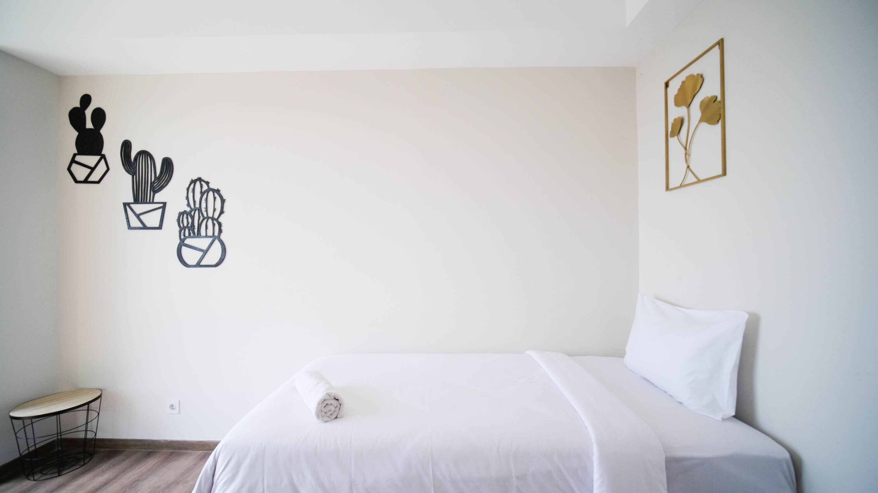 A minimalist hotel room with a neatly made bed, a small round table, and wall art depicting cacti.