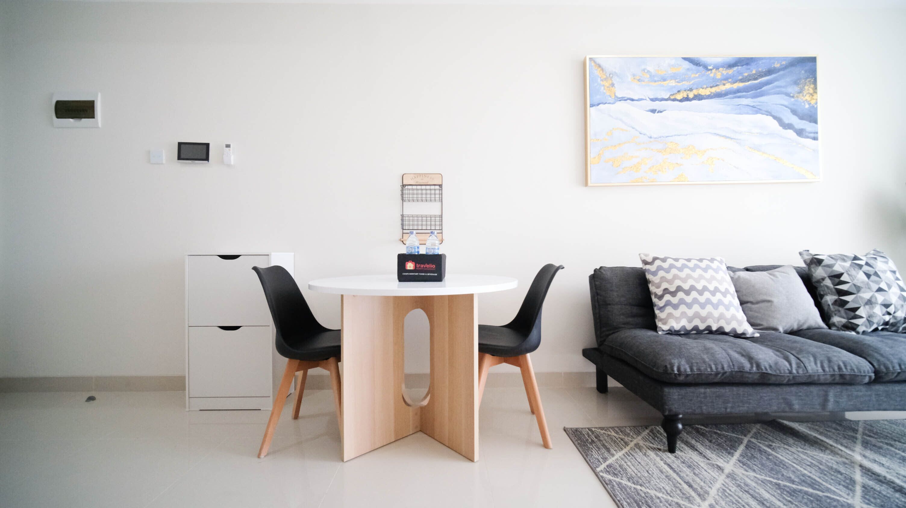 A modern living room with a round dining table, two black chairs, a grey sofa with patterned cushions, a travelio branded water bottle holder, and a large abstract painting on the wall.