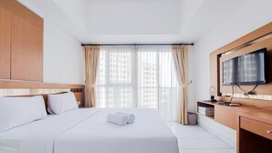 A hotel room with a large bed, a wooden headboard, a TV mounted on the wall, and a window with curtains. - Warm And Enjoy Living Studio Casa De Parco Apartment (South Tangerang)