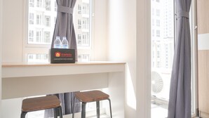 Ein Hotelzimmer mit Fenster, Ablage mit Wasserflaschen, Schreibtisch und zwei Hocker.