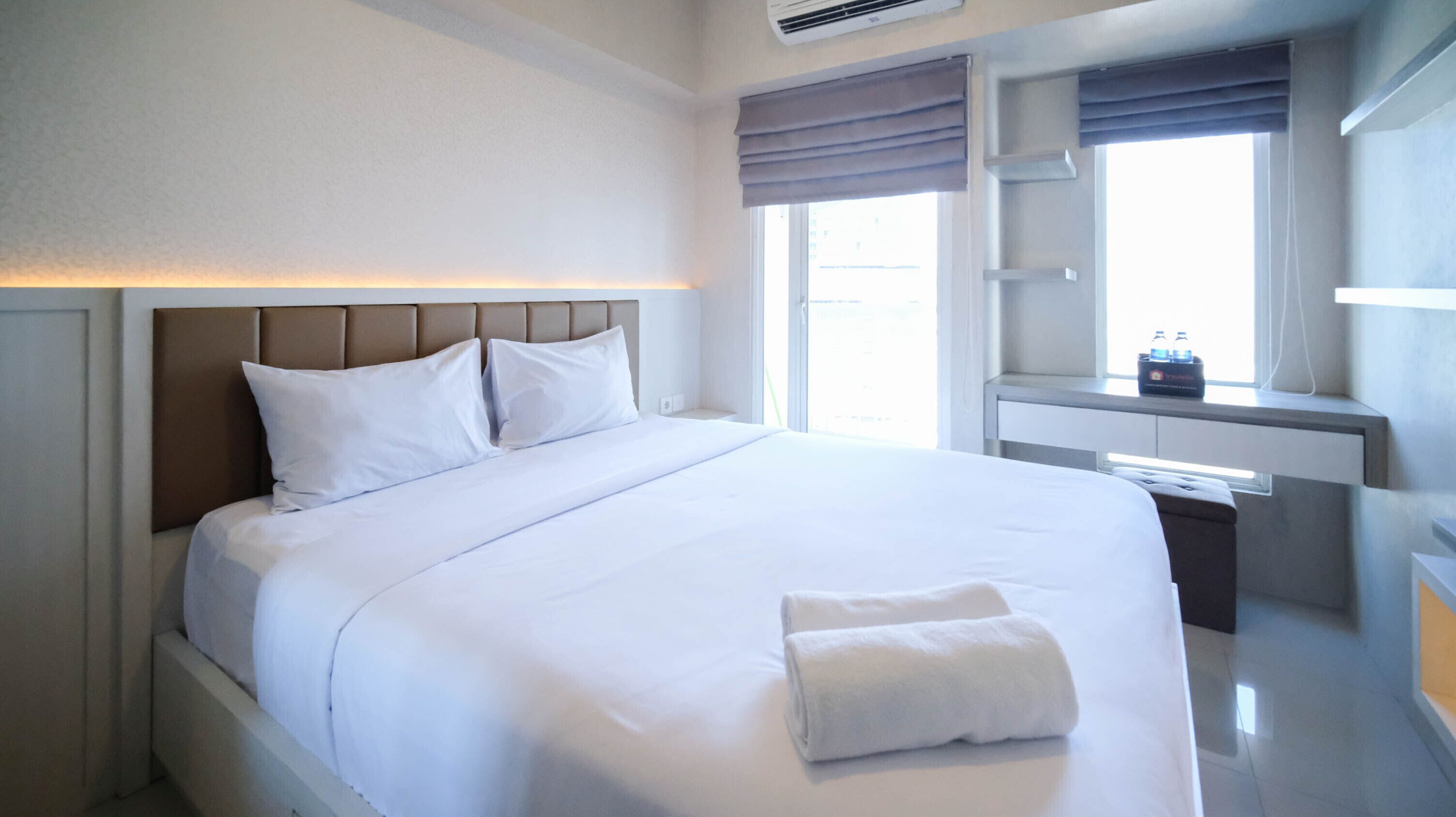 A neatly made bed with white linens and pillows, a bedside table with a bottle, and a window with curtains.