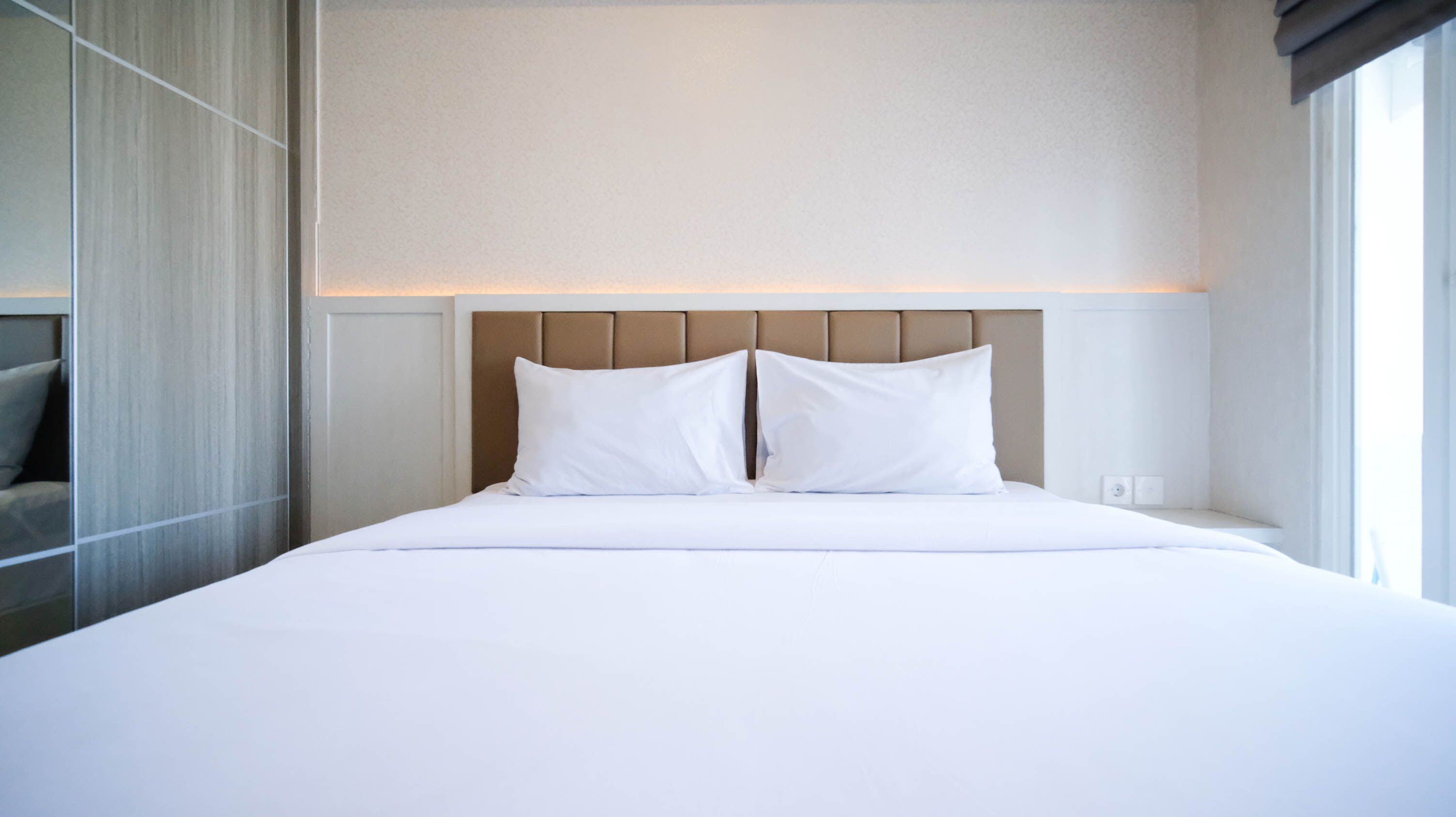 A modern hotel room with a large bed, a headboard with a light panel, and a view of the city through the window.
