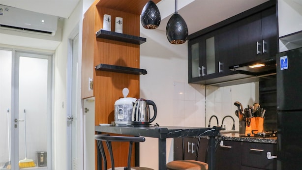 A compact kitchen with a black countertop, two bar stools, a coffee maker, and a black refrigerator. - Warm And Homey 2Br At Sentraland Cengkareng Apartment (Jakarta)