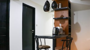 A small dining area with a black table and two chairs, a wooden shelf with kitchen items, and a black door. - Warm And Homey 2Br At Sentraland Cengkareng Apartment (Jakarta)