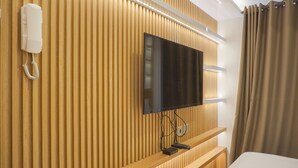 A hotel room with a wooden panel wall, a mounted flat-screen TV, a bedside telephone, and a bedside table with a remote control. - Homey Living Studio Room At Tokyo Riverside Pik 2 Apartment (Teluknaga)