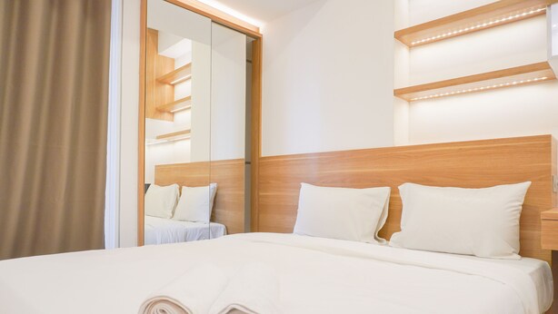 A neatly made bed with white linens and towels, a wooden headboard, and a mirror reflecting a staircase. - Homey Living Studio Room At Tokyo Riverside Pik 2 Apartment (Teluknaga)