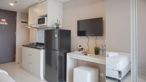 Eine kompakte Küche mit Kühlschrank, Mikrowelle und Herd. Ein kleiner Tisch mit Pflanze und Wasserflaschen steht dort. Ein Bett ist im angrenzenden Raum zu sehen.