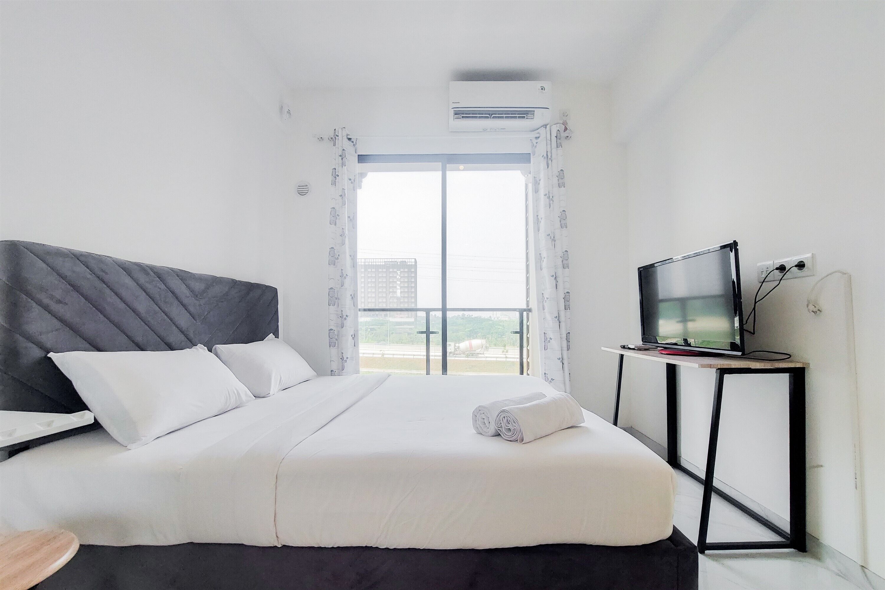 A hotel room with a bed, a TV on a stand, and a view of the city through the window.