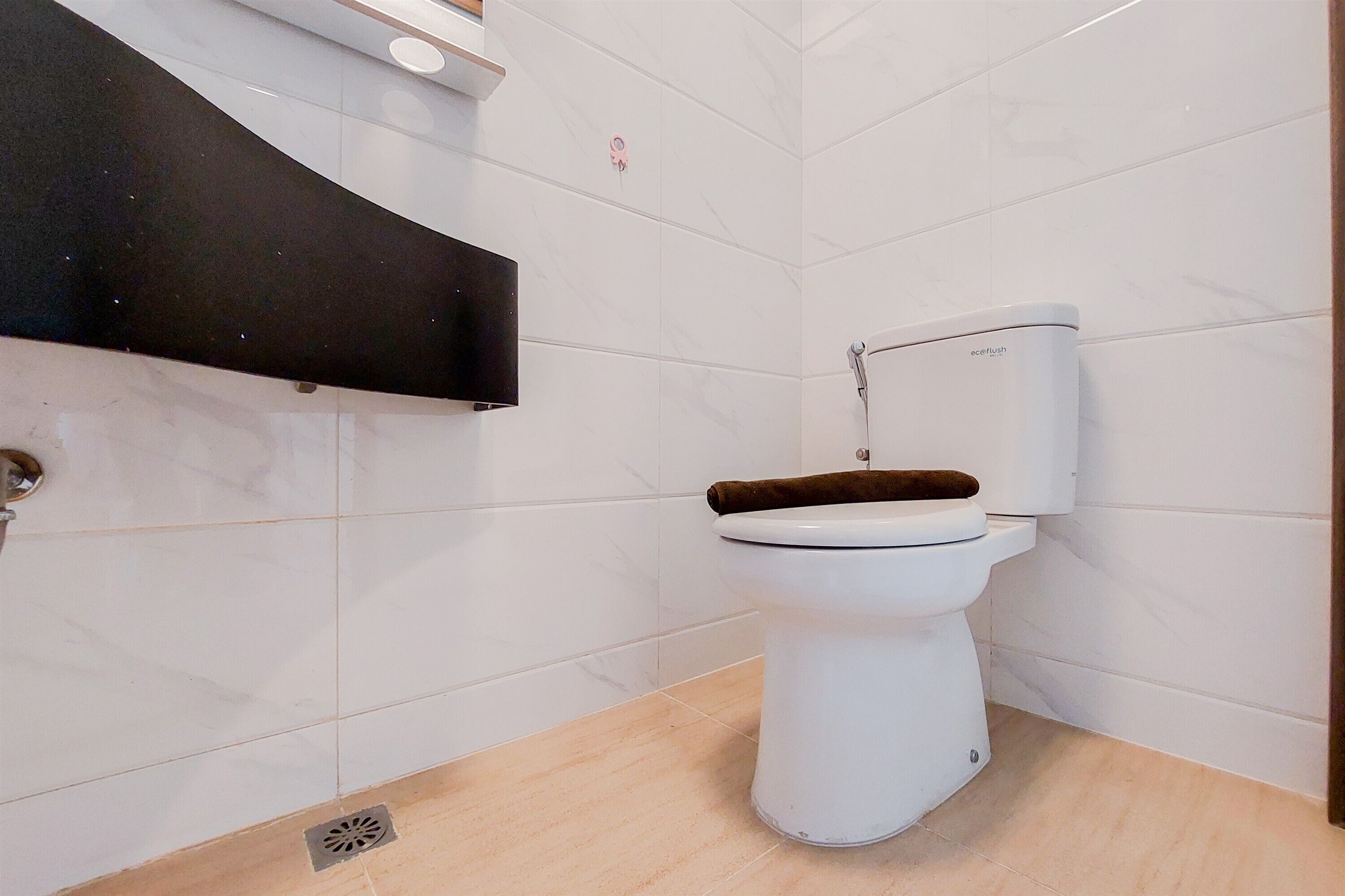 Ein modernes Badezimmer mit weißen Fliesen, einer Toilette mit braunem Sitz und einem schwarzen Regal darüber.