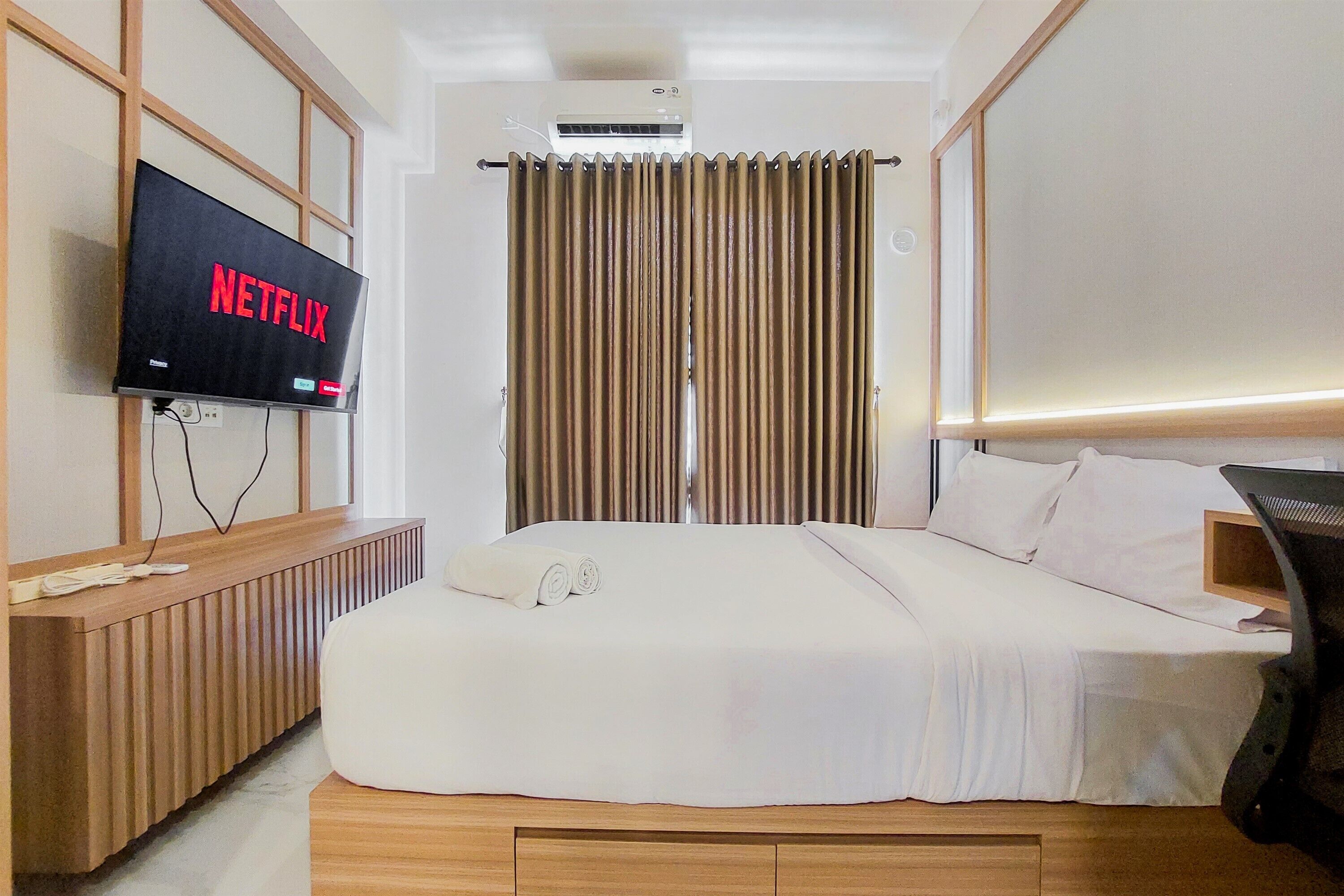 A hotel room with a bed, a TV displaying 'NETFLIX', a wooden bedside table, and a window with curtains.
