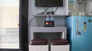 A modern room with a flat-screen TV mounted above a white console table holding two bottles of water and a travelio branded box. A blue refrigerator with magnets and a glass shelf is visible to the right. - Compact And Elegant Studio Maple Park Sunter Apartment (Jakarta)