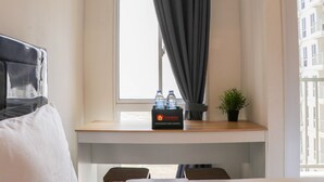 A hotel room with a bed, a window with curtains, a small table with a box of water bottles, and a potted plant. - Brand New Studio At Tokyo Riverside Pik 2 Apartment (Teluknaga)
