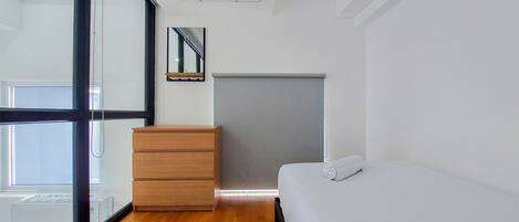 Un dormitorio moderno con un aparador de madera, una cama, un mueble gris y un amplio ventanal con persianas.