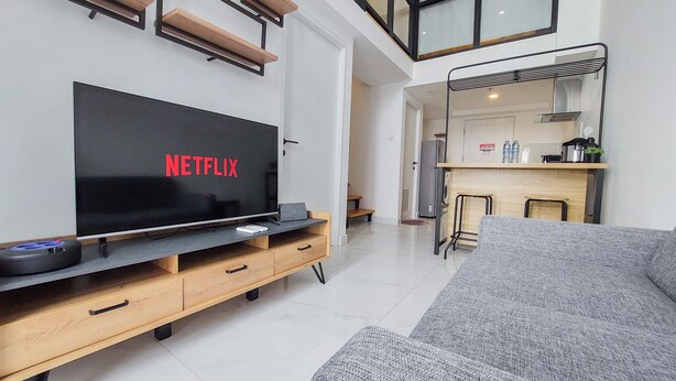 Una sala de estar moderna con televisor de pantalla plana, mueble de madera para entretenimiento, sofá gris y una zona de cocina al fondo.