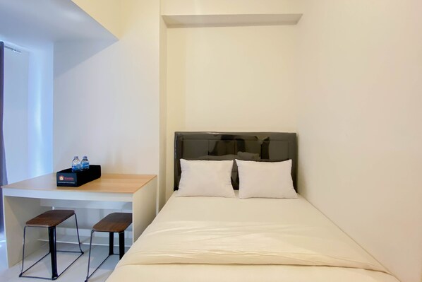 A small hotel room with a bed, two white pillows, a wooden desk with a chair, and a small table with two bottles.