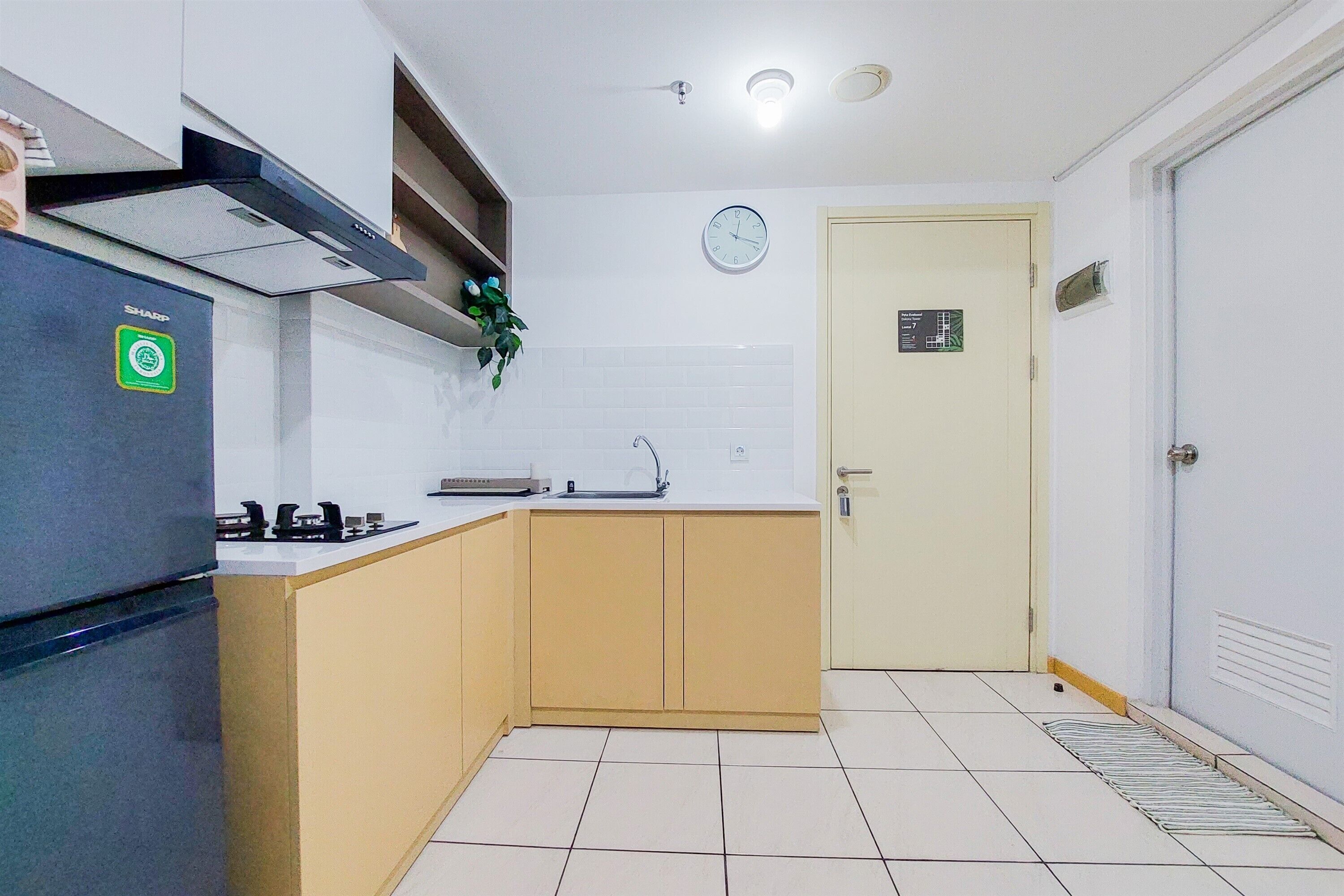 A compact kitchen with a refrigerator, stove, and sink, a clock on the wall, and a door with a sign.