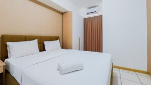 Una habitación de hotel con una cama, ropa de cama blanca, cabecera de madera, mesita de noche, ventana con cortinas y un equipo de aire acondicionado.
