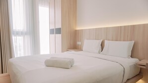 A neatly made bed with white linens and a rolled towel, a wooden headboard, and a bedside table with a lamp. - Homey And Warm Studio Amazana Serpong Apartment (South Tangerang)