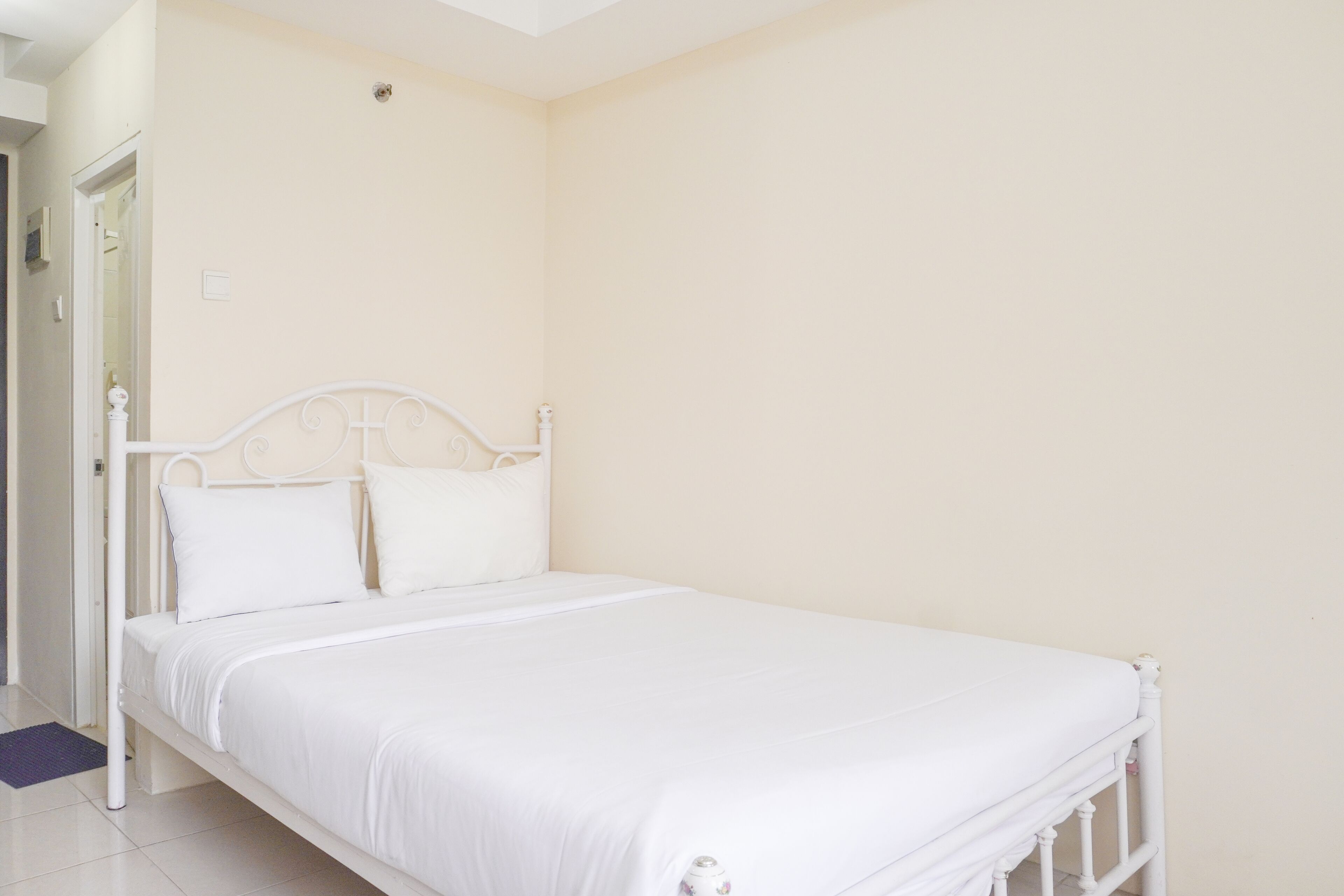 Un lit en métal blanc dans une pièce aux murs de couleur crème, avec une porte sur le côté gauche.