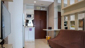 A modern hotel room with a flat-screen TV, a covered sofa, a small table with a clock, and a kitchen area visible through an open door.