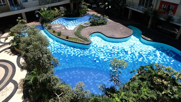 Une piscine sur le toit, dont le fond présente un motif à carreaux, entourée de verdure et bordée d’un immeuble à plusieurs étages doté de balcons.