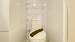 A toilet with a brown towel on the seat, white tiles on the walls, and a toilet paper holder. - Warm And Simply Look Studio Apartment At Tokyo Riverside Pik 2 (Teluknaga)