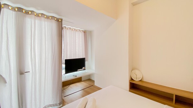 A hotel room with a bed, a TV on a stand, a wooden shelf, and a window with curtains. - Warm And Simply Look Studio Apartment At Tokyo Riverside Pik 2 (Teluknaga)